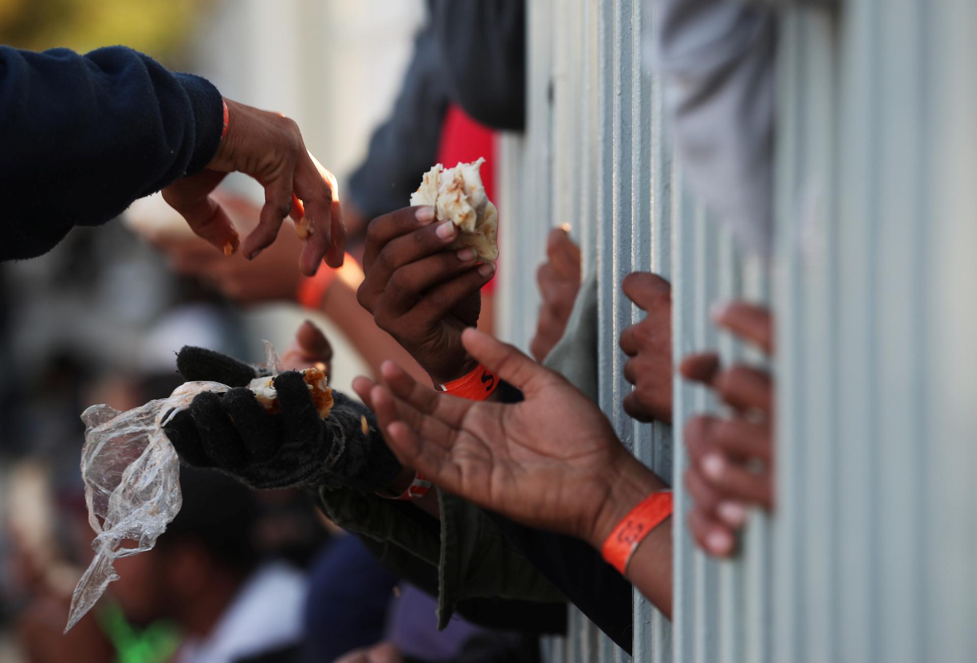 migrants reach through a fence with pieces of food