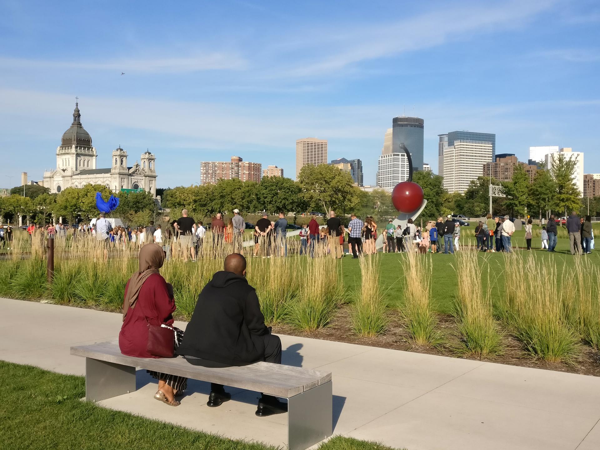 Couple sits on bench facing large statue of cherry in spoon, other milling around on grass