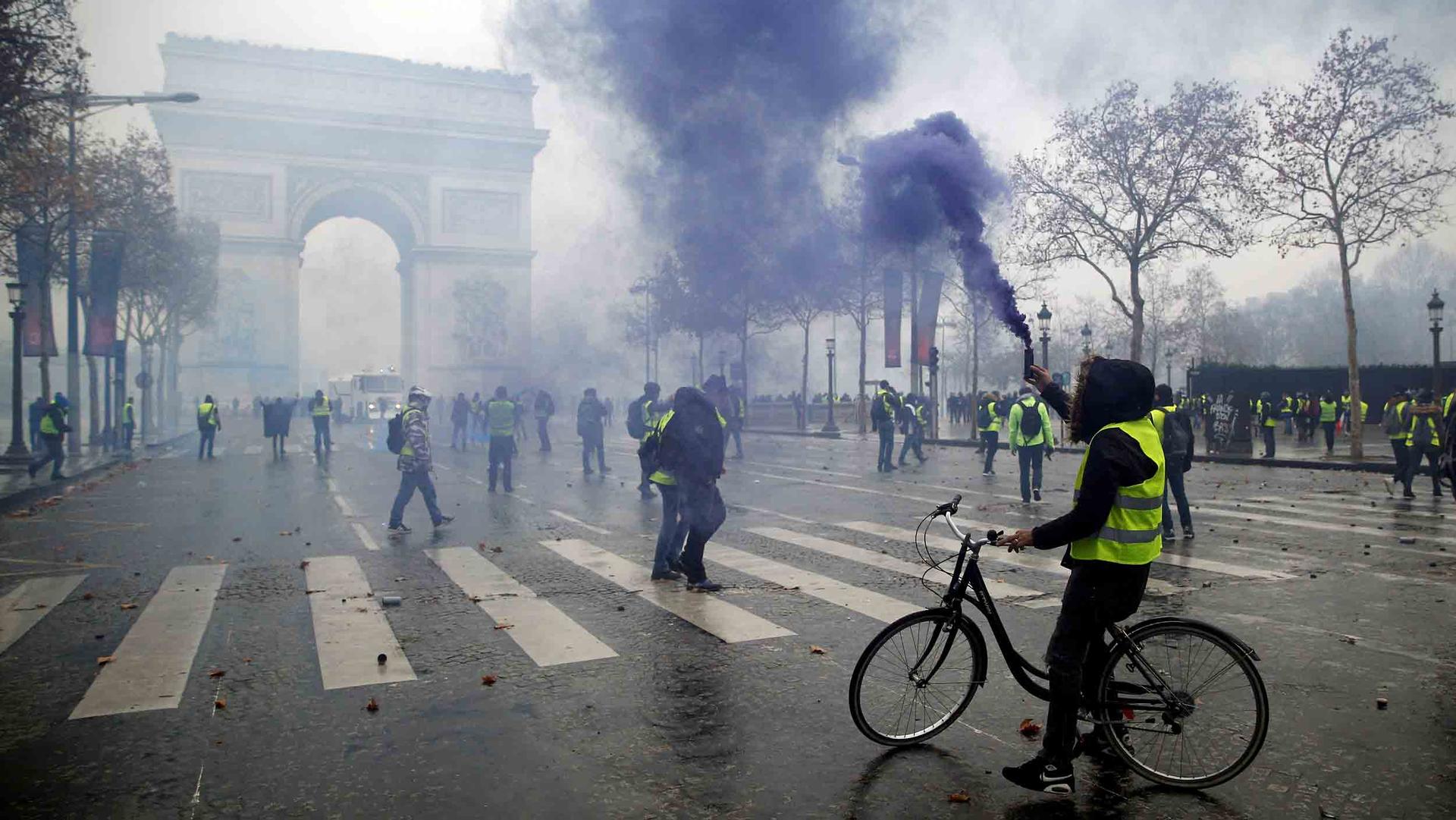 A man stands on a bicycle with a smoke flare in his hand, wearing a bright yellow vest in the street before the Arc de Triomphe. About 50 others also wearing yellow vests are also in the street.