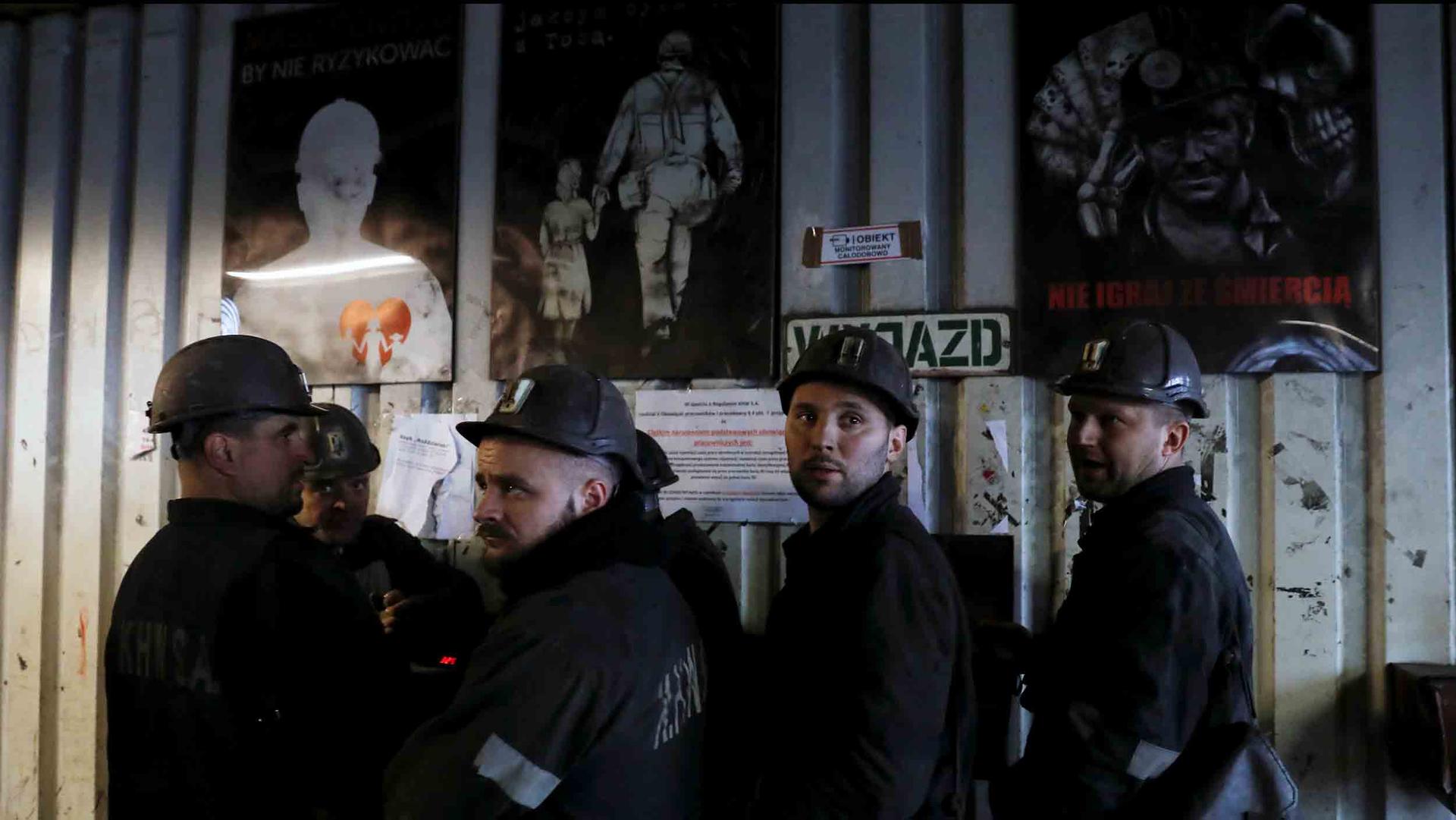 A group of men wearing hard hats and dark grey clothes stand in front of posters depicting miners at work