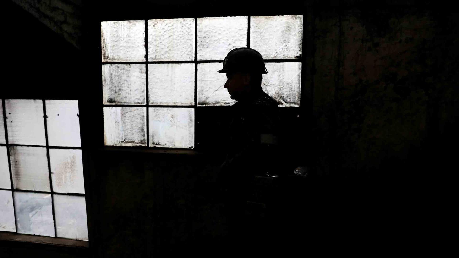 A man wearing a hard hat is silhouetted against old heavy windowpanes in a factory.