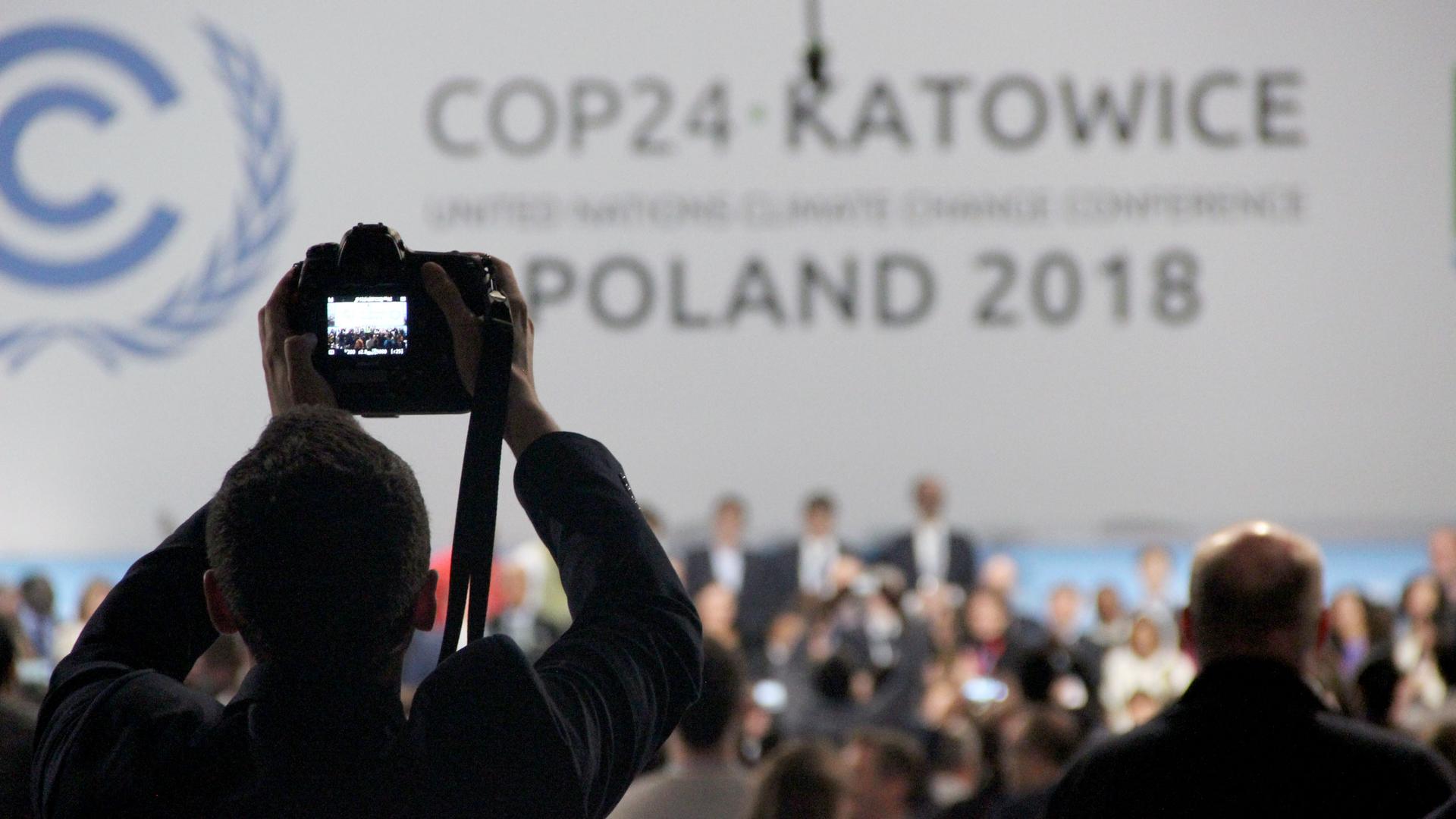 A digital camera is held up over the heads of others and the screen lights up as it takes a photo of those assembled on the stage at the front of the room