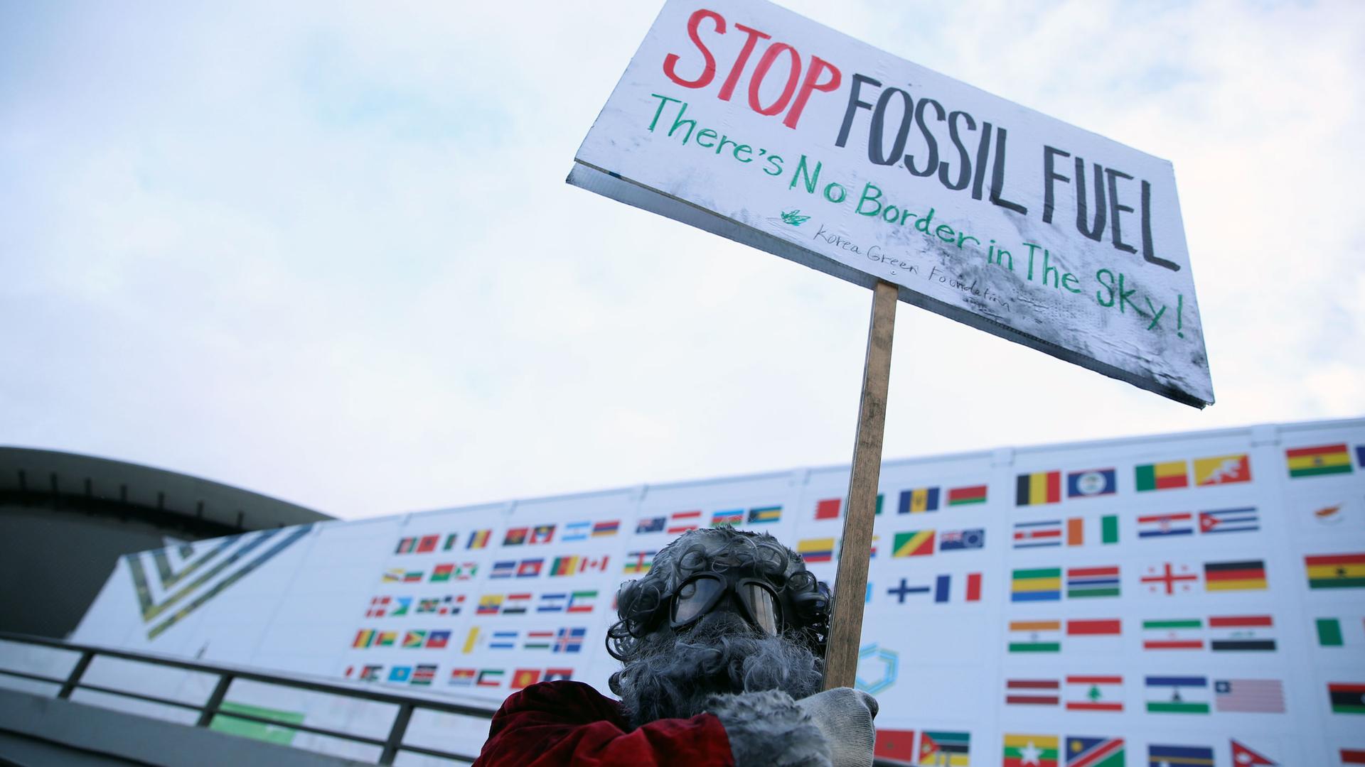 A man wearing googles holds up a sign that says "stop fossil fuel! there is no border in the sky!"