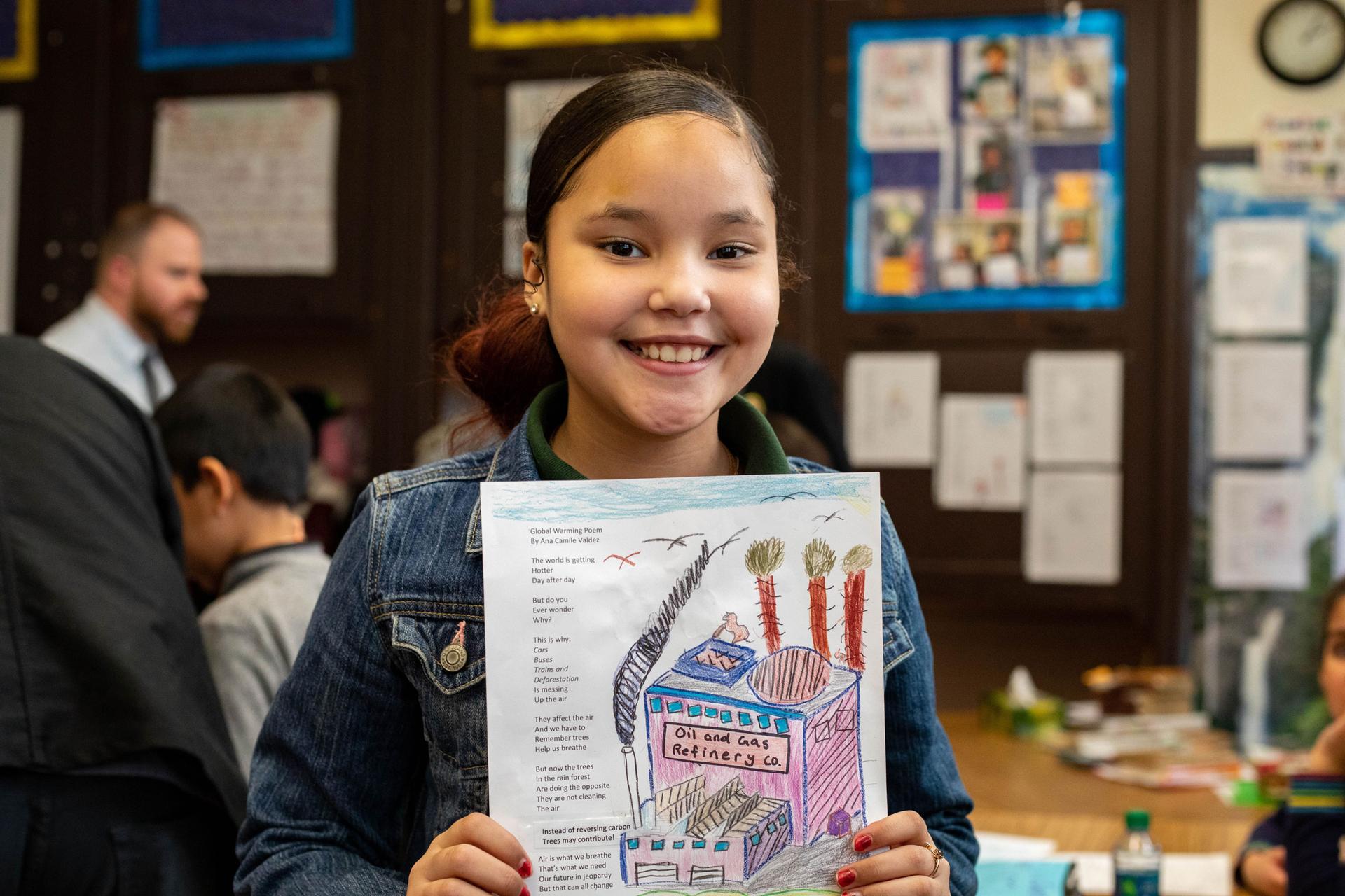A young girl holds a photo of her poem with her illustrations on it in her school classroom and smiles at the camera
