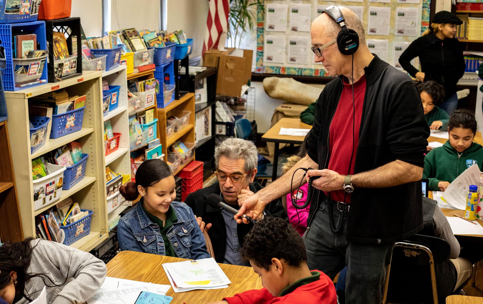 A young girl sits at a school table and reads while a man holds a microphone next to her and records her reading.