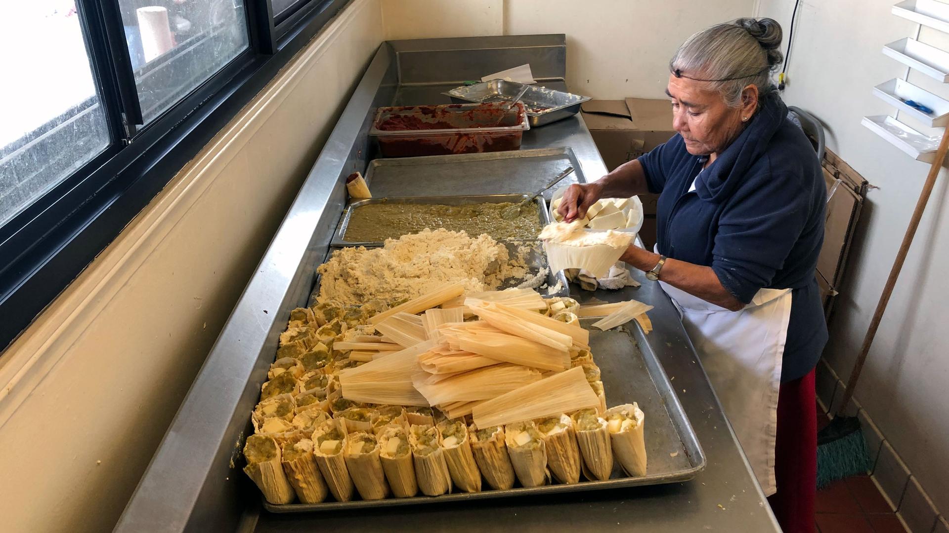 A woman stands a at a table assembling tamales