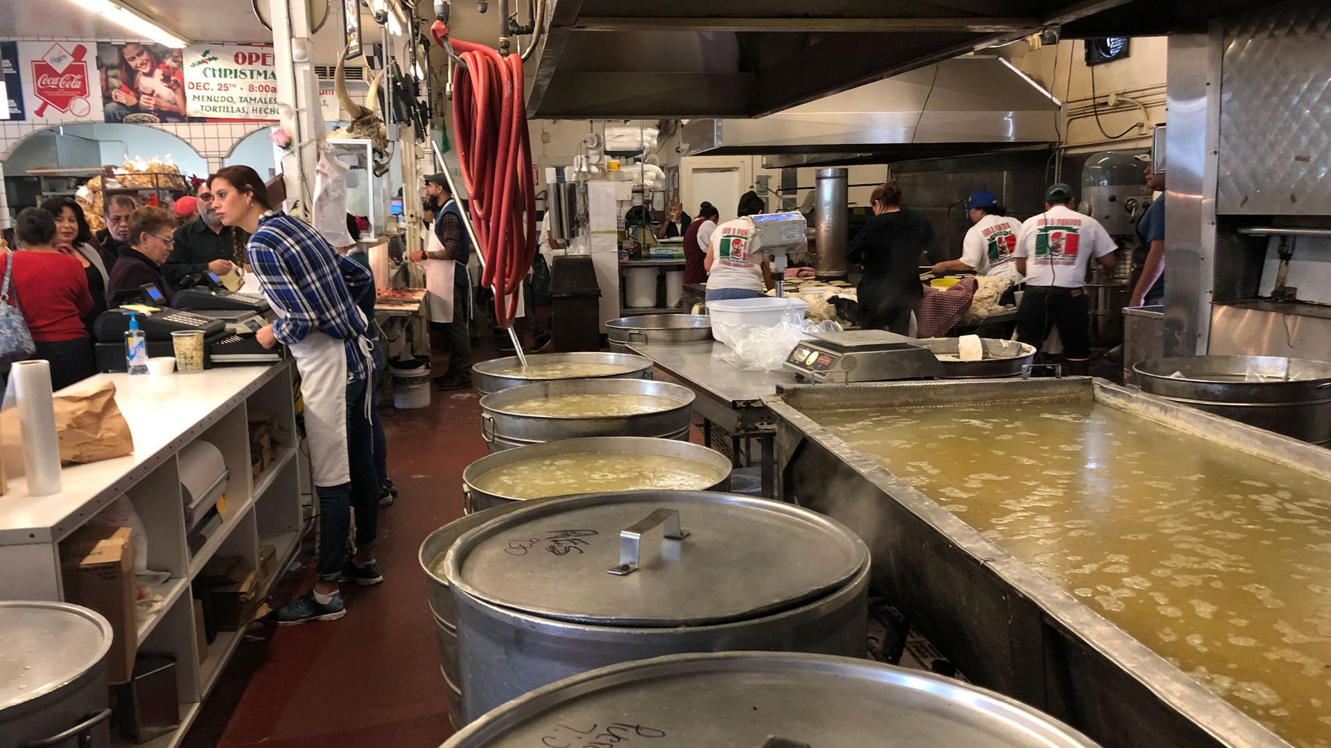 At a counter in a Latino market, customers wait to buy food while workers cook in the back. In the foreground are huge steel pots. 