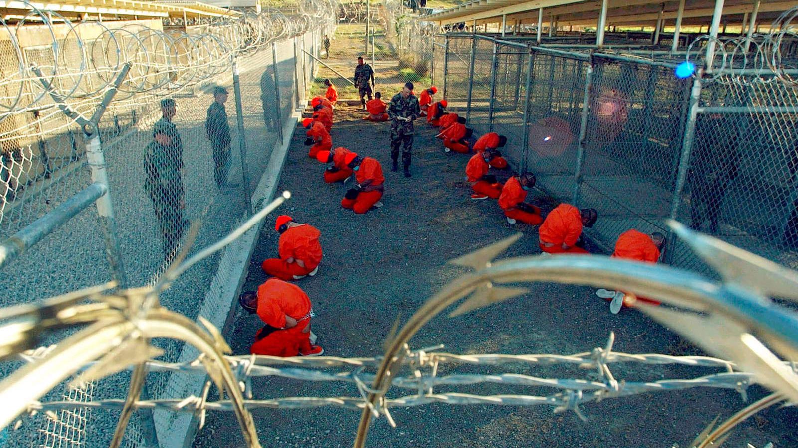 Detainees wearing orange jumpsuits crouch down within a barbed wire protected area. 