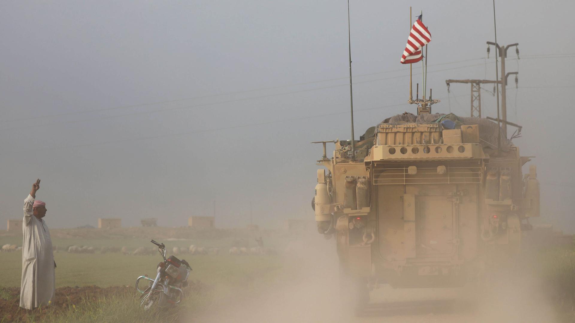 A man gestures at US military vehicles as dust colors the air