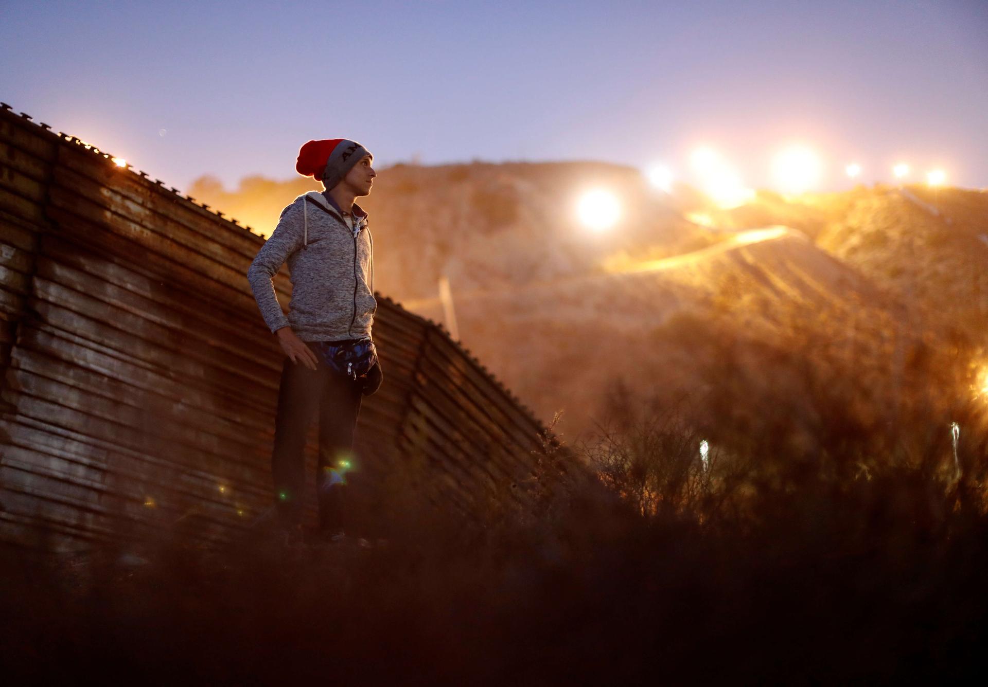 A migrant from Honduras stands in front of the border wall with the sun shining behind him 