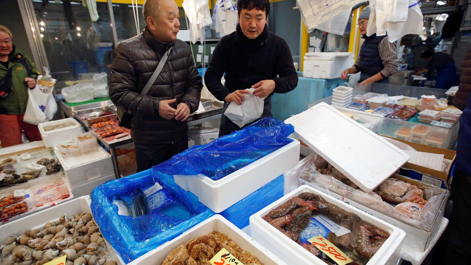 Two men stand near fish in bins at fish market.