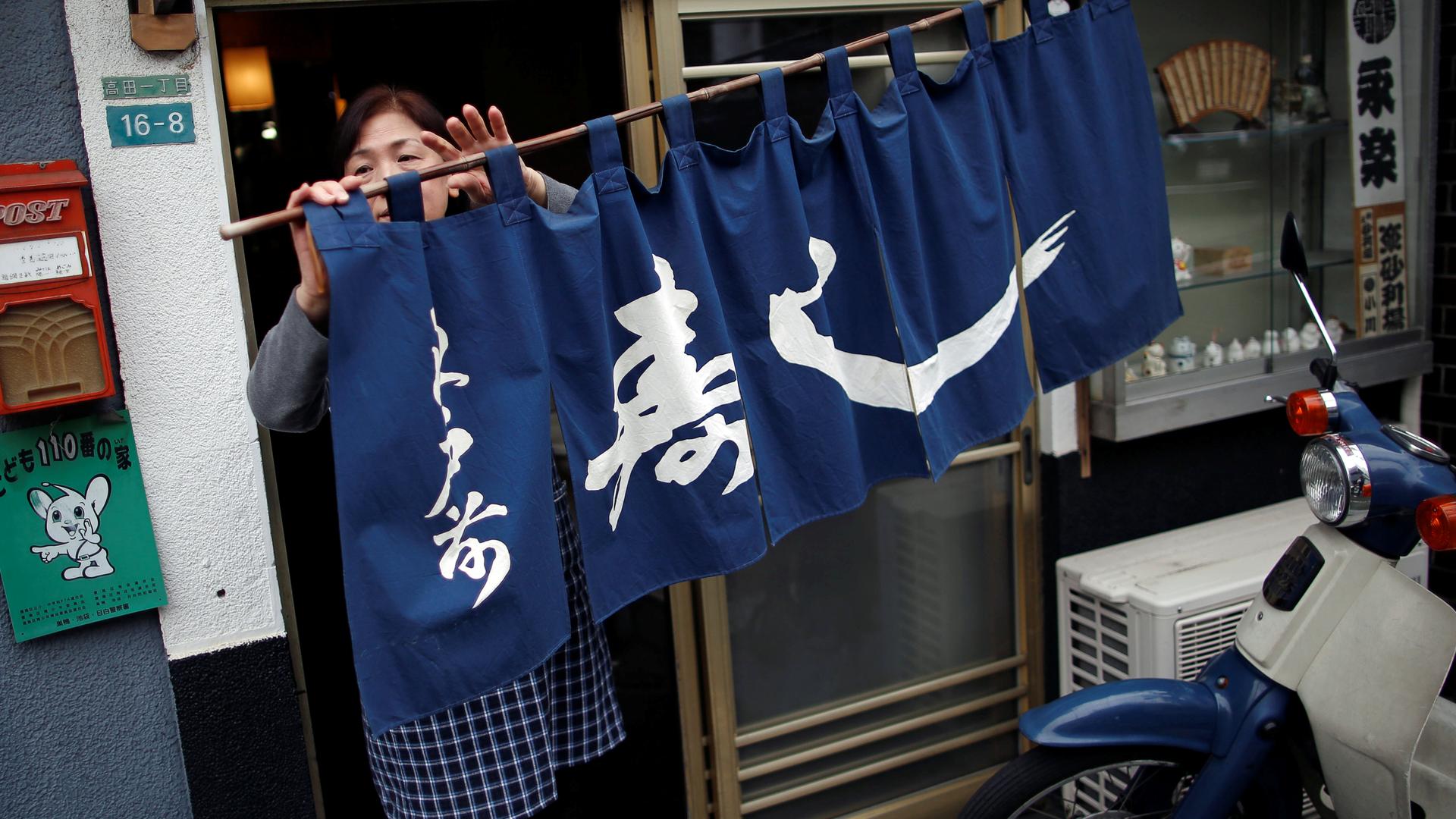 A woman moves a blue curtain with Japanese script as she opens the restaurant.