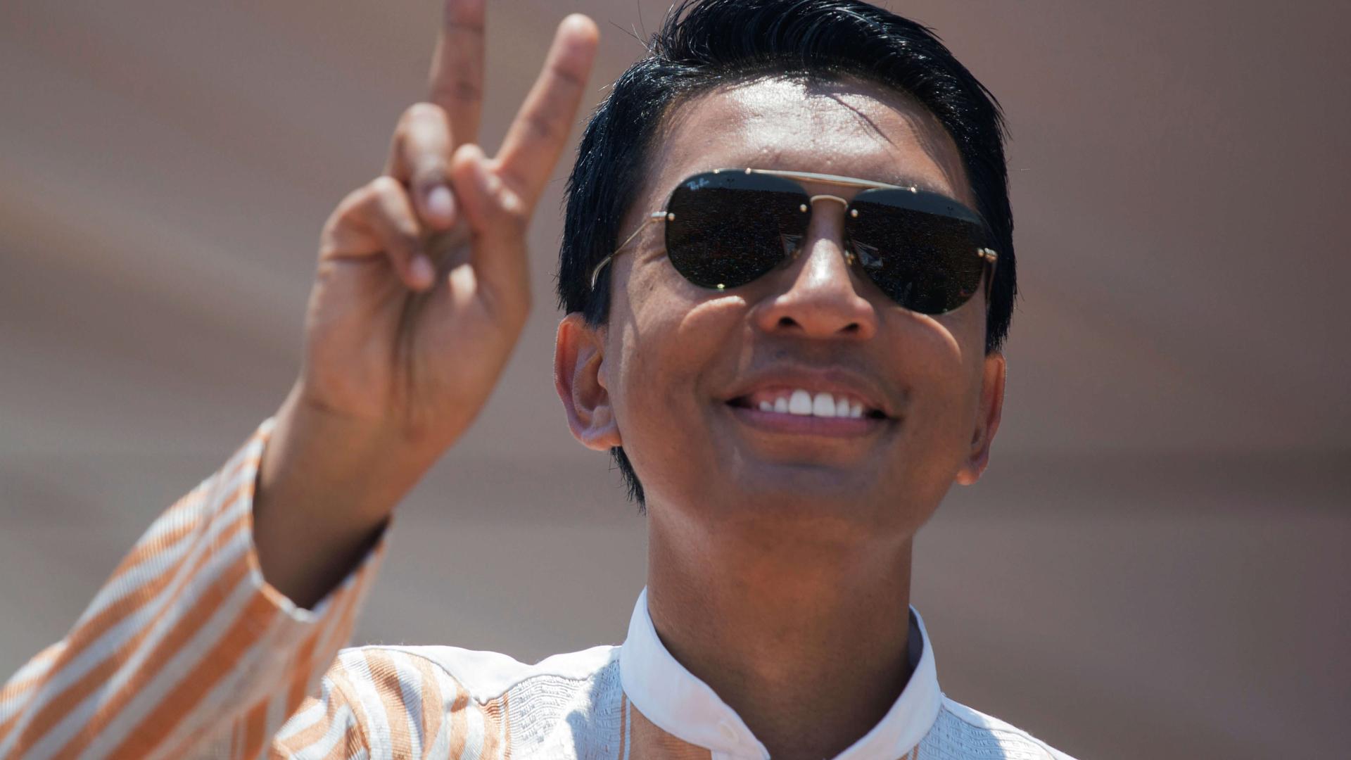 A man with glistening black hair wearing sunglasses waves to the crowd. 