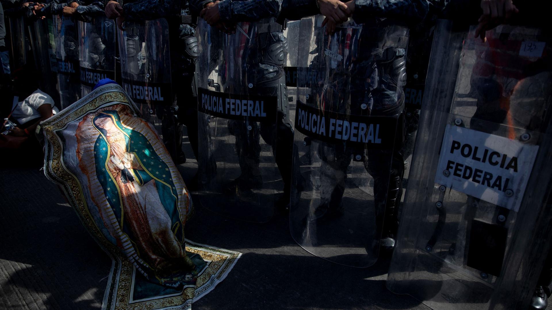 A migrant is shown wrapped with a banner depicting the Virgin of Guadalupe in front of a riot police with shield in Tijuana, Mexico.