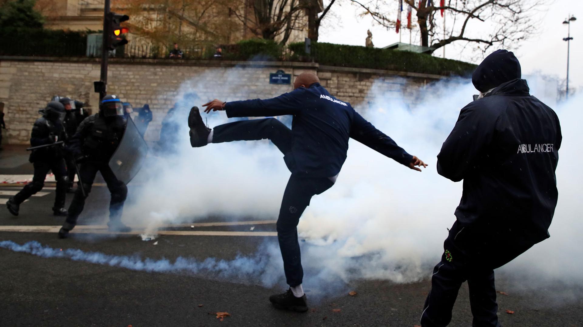 An ambulance driver in France is shown kicking a can of smoke with French riot police in the background.
