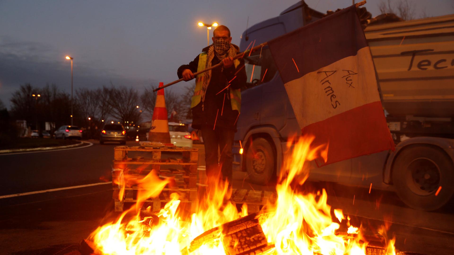 A protester wearing a yellow vest is shown holding a flag near burning debris at the approach to the A2 Paris-Brussels highway, Dec. 4, 2018.