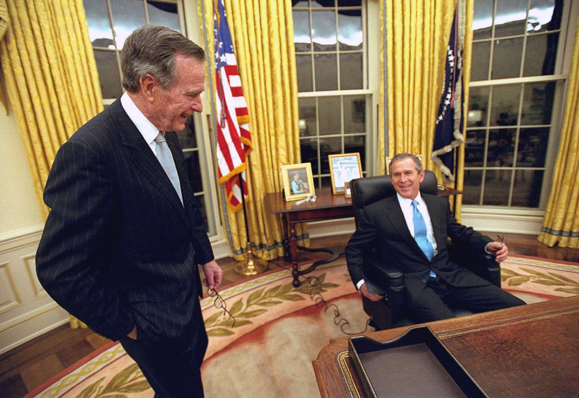 George W. Bush sits at the desk in the Oval Office while his father, H.W. Bush, stands off to the left.