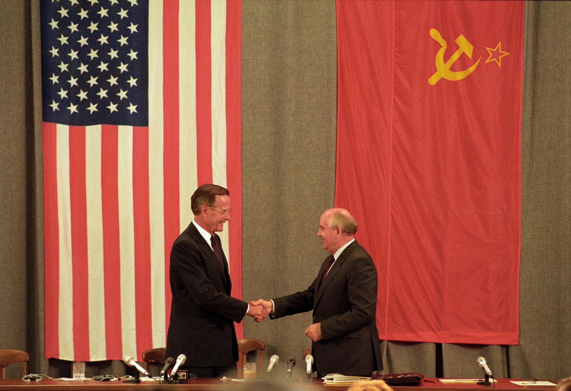 President HW Bush stands in front of an American flag and Soviet President Mikhail Gorbachev stands in front a Soviet flag as the two shake hands.