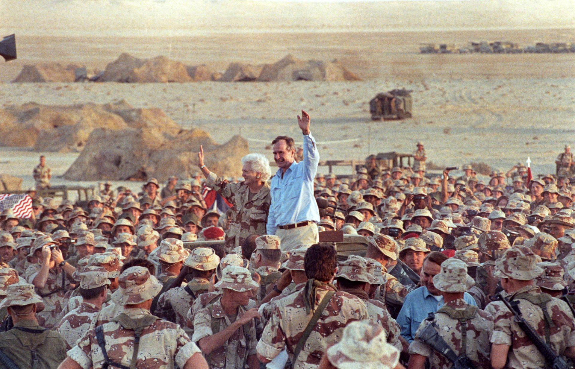 President HW Bush and his wife wave while surrounded by a large crowd of soldiers wearing tan and khaki fatigues