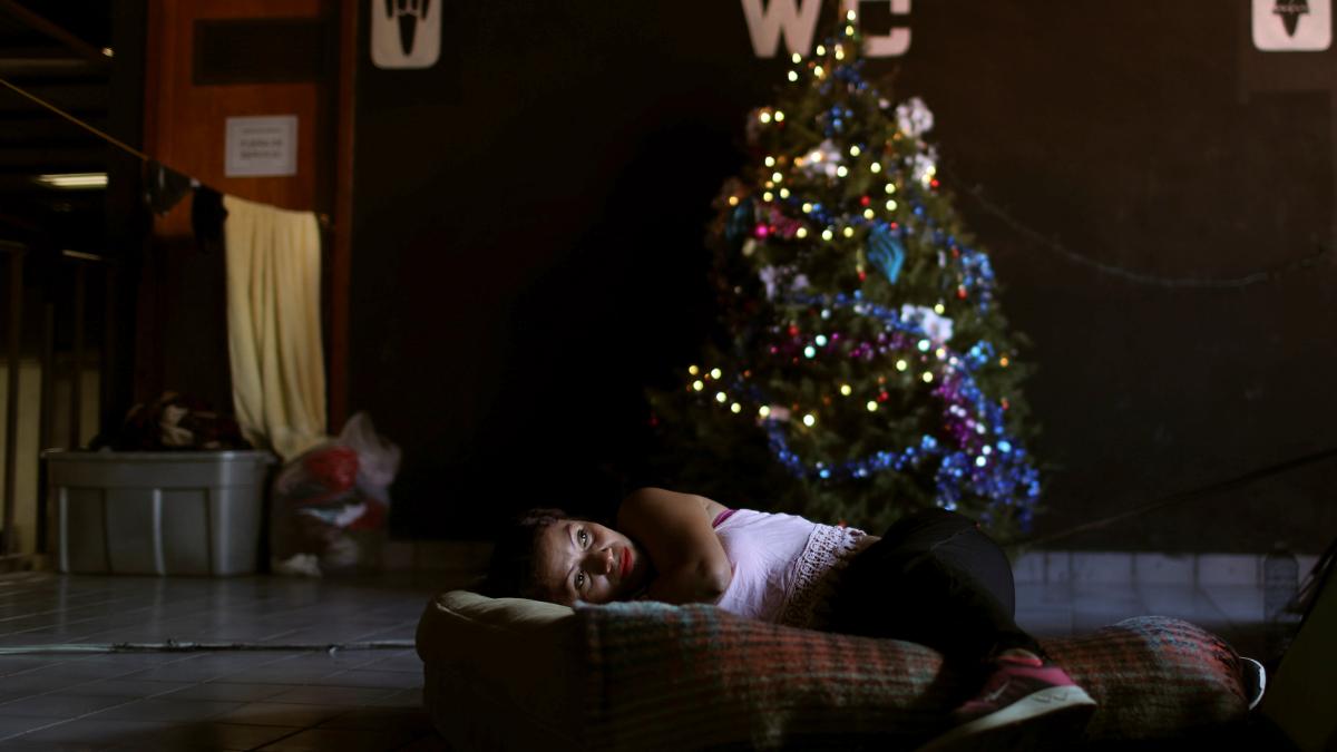A woman lays on a mattress on the floor in front of a Christmas tree in a dark room in a shelter in Tijuana, Mexico.