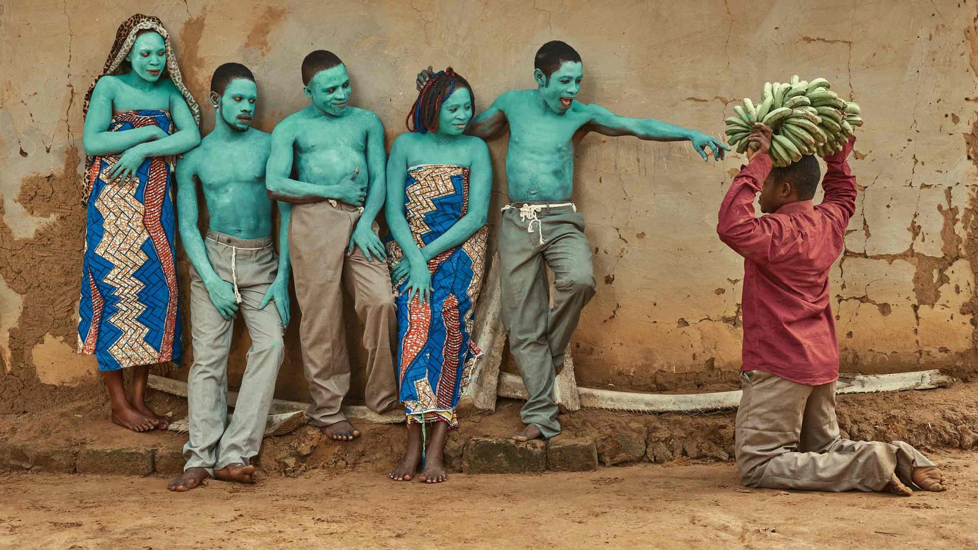 Five people are painted with a light green paint and post together. Another man is on his knees with bananas. 