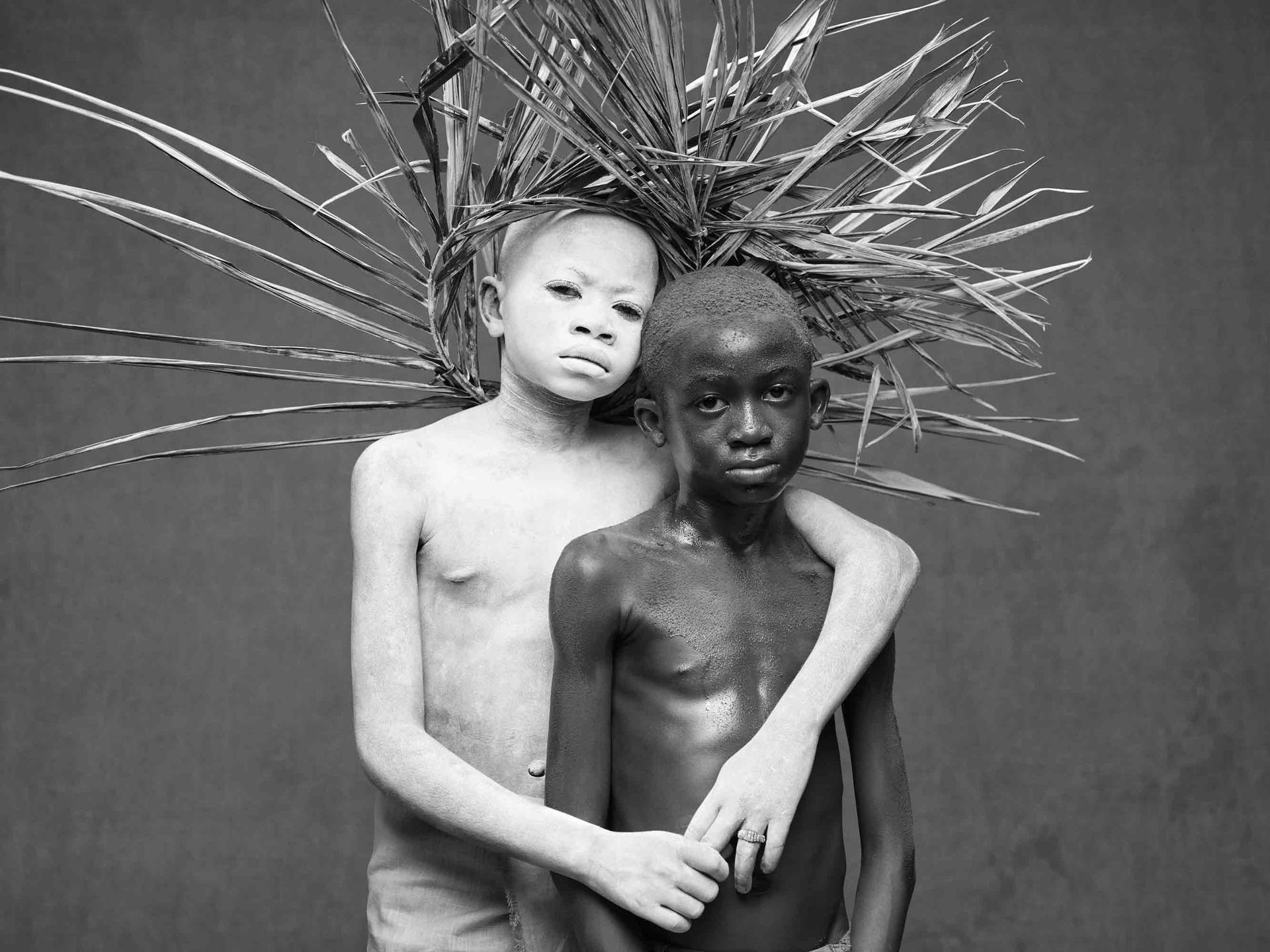 A boy wearing white body paint and a wooden headdress has an arm around another boy as they pose for a portrait