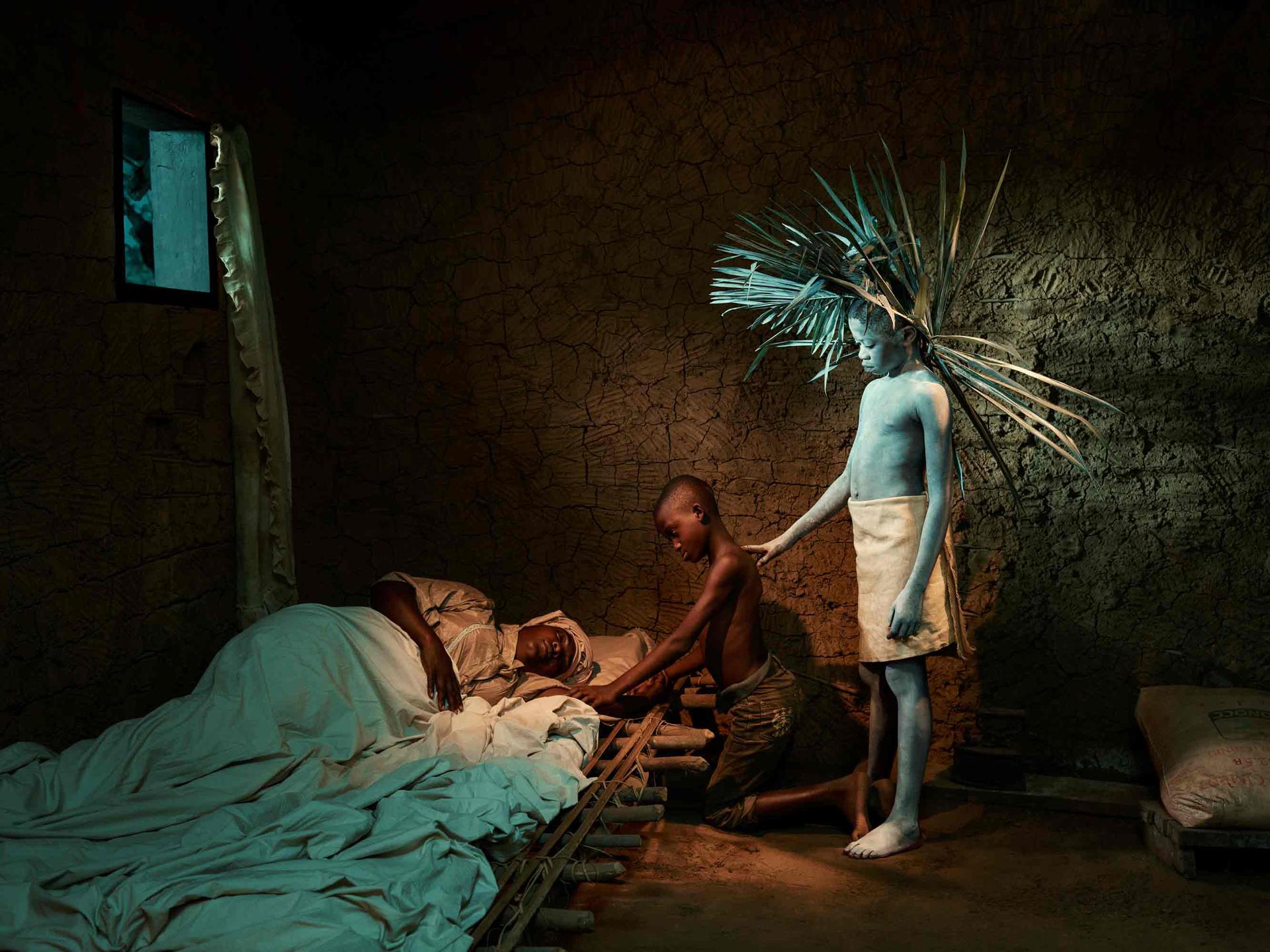 A boy painted white and wearing a headdress of wooden sticks stands over a person lying in bedding on the floor