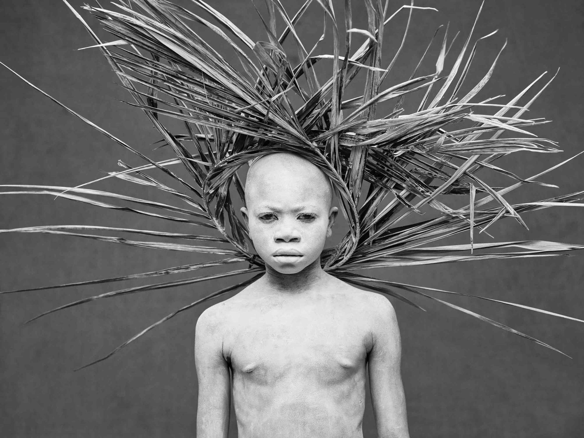 A boy whose body is painted white wears a crown of wooden sticks that looks like a sunburst behind his head poses for a photo