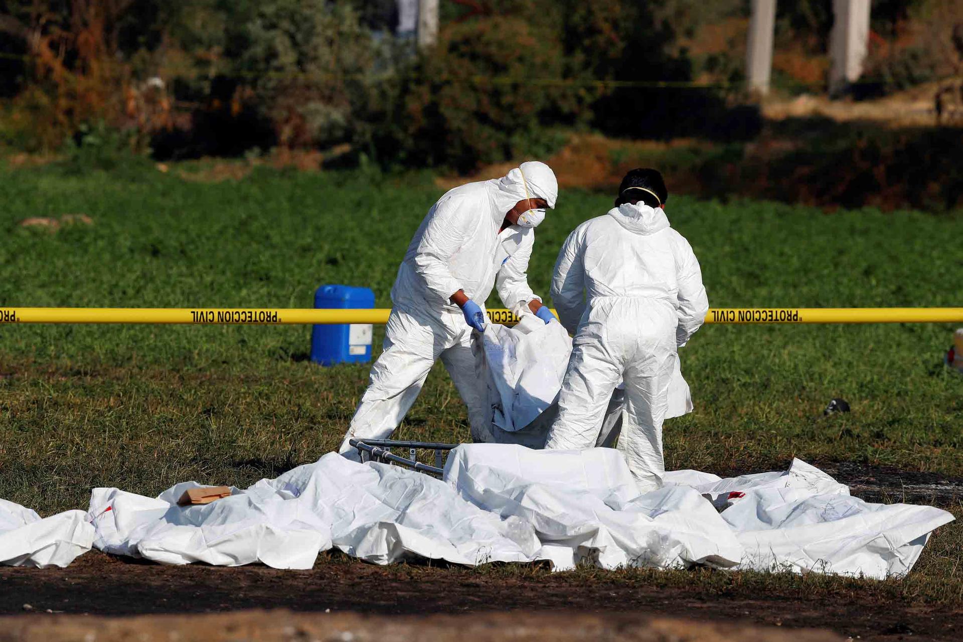 Men in white suits arrange bodies in white bags behind a yellow tape line