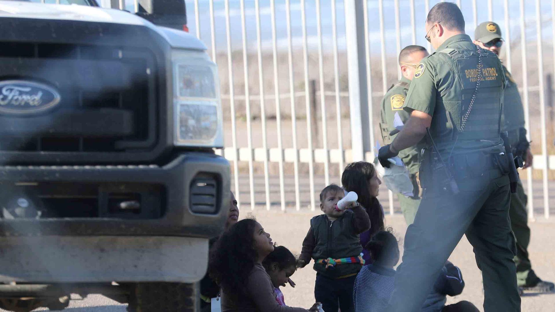 A migrant woman and her children sit beside a truck while three border patrol officers take them into custody