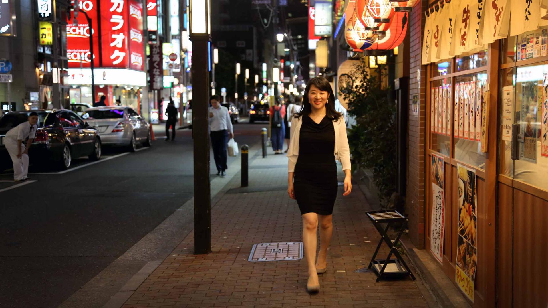 A woman in a black dress stands on a city street in Japan. Signs with Japanese characters are behind her, on restaurants and billboards.