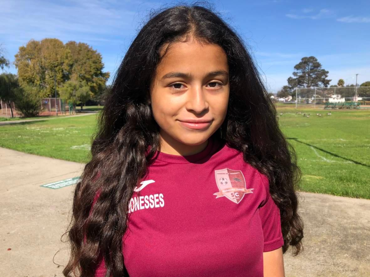 A headshot of Bay Area teen Crista Ramos, who is suing the federal government. She wears a red shirt, has long dark hair, and looks directly into the camera.