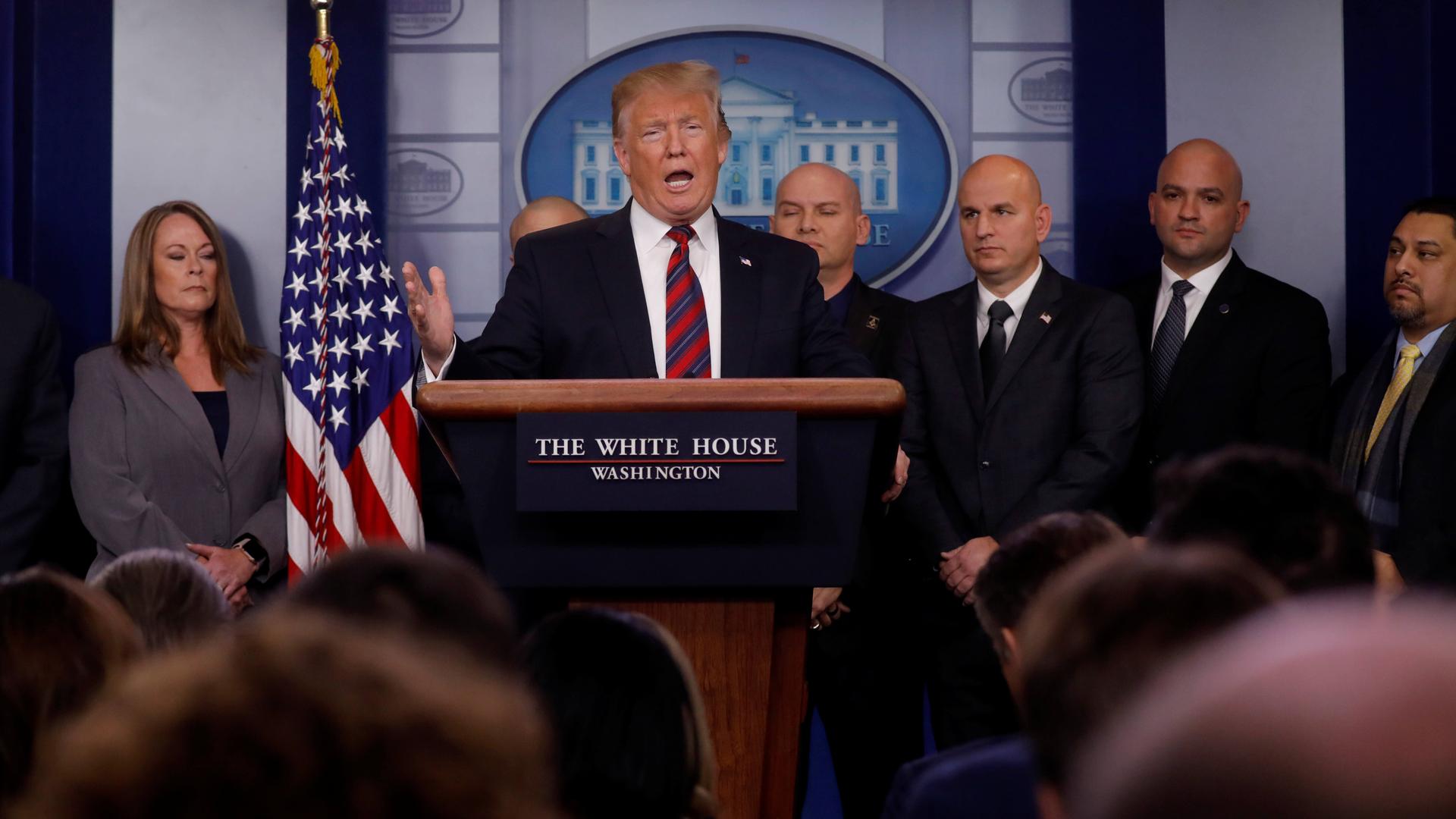 President Trump speaks at a podium while 3 bald men stand behind him.