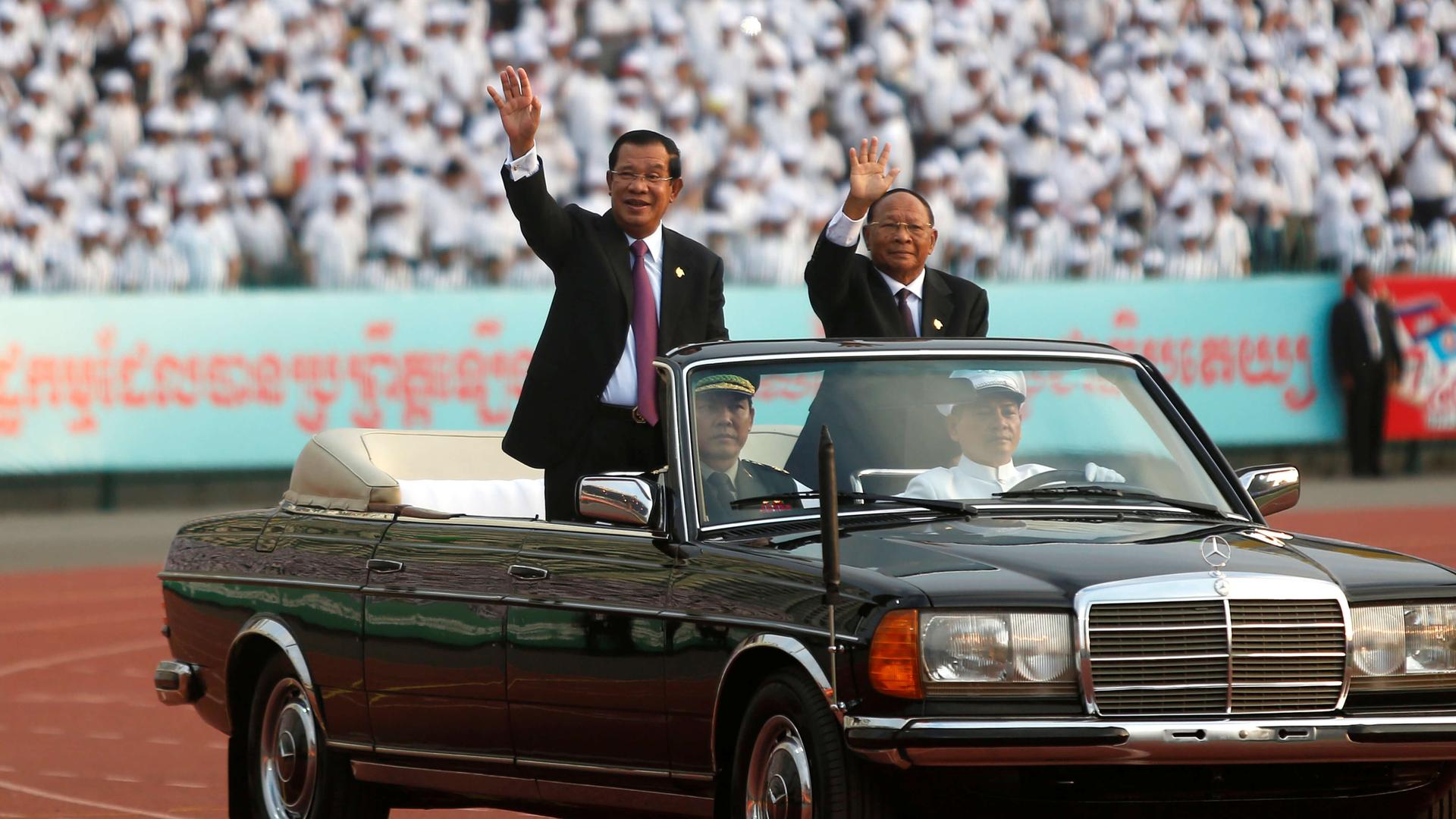 Two men wave from a convertible car.