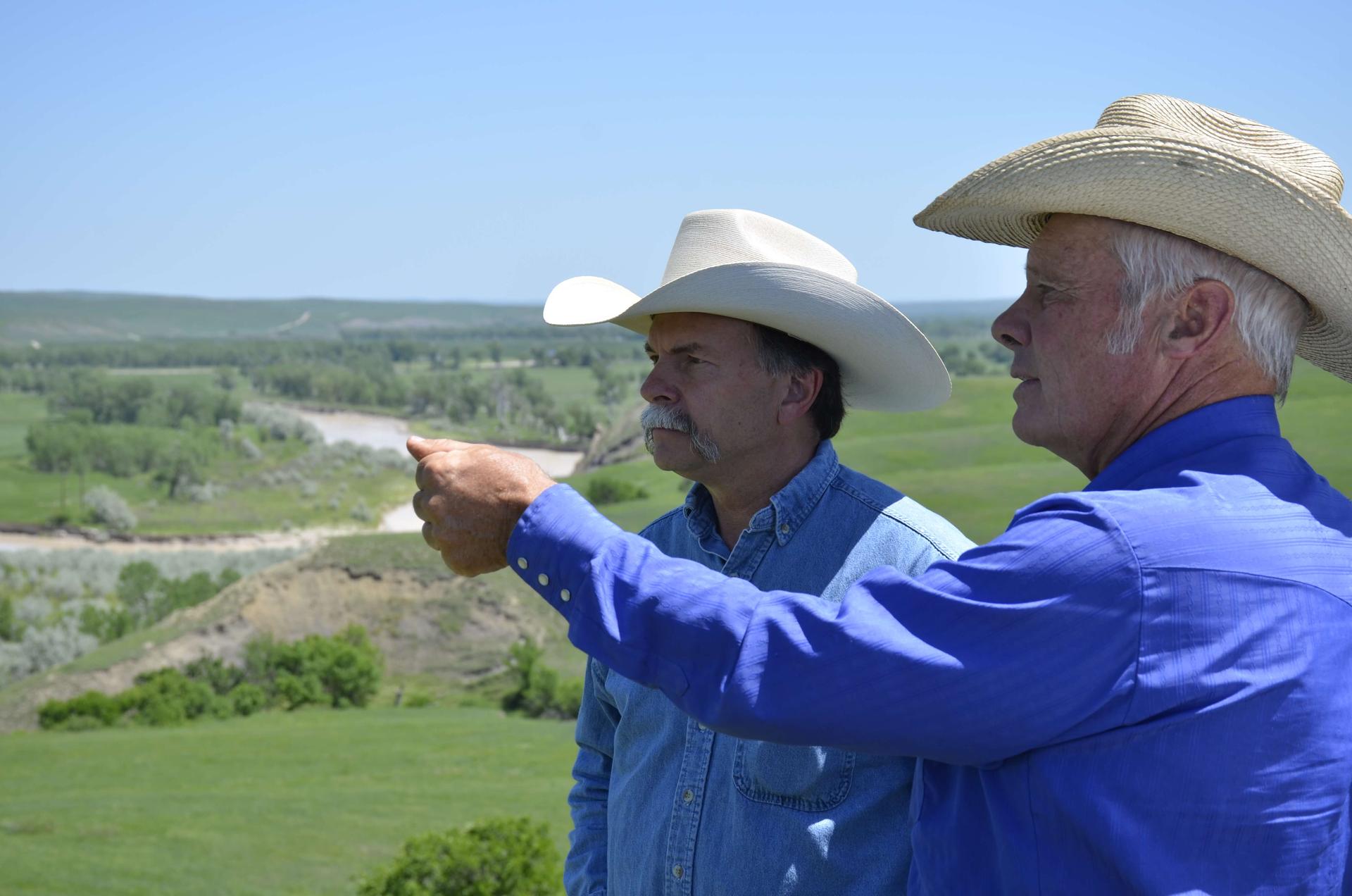 two men wearing cowboy hats stand on a ranch and point