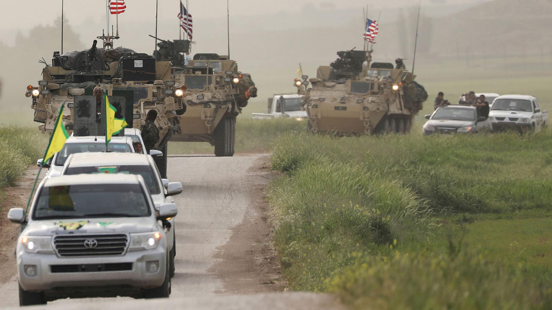 Kurdish fighters driving Toyota SUVs with green and yellow flags on top are shown driving in head of US military MRAP vehicles.