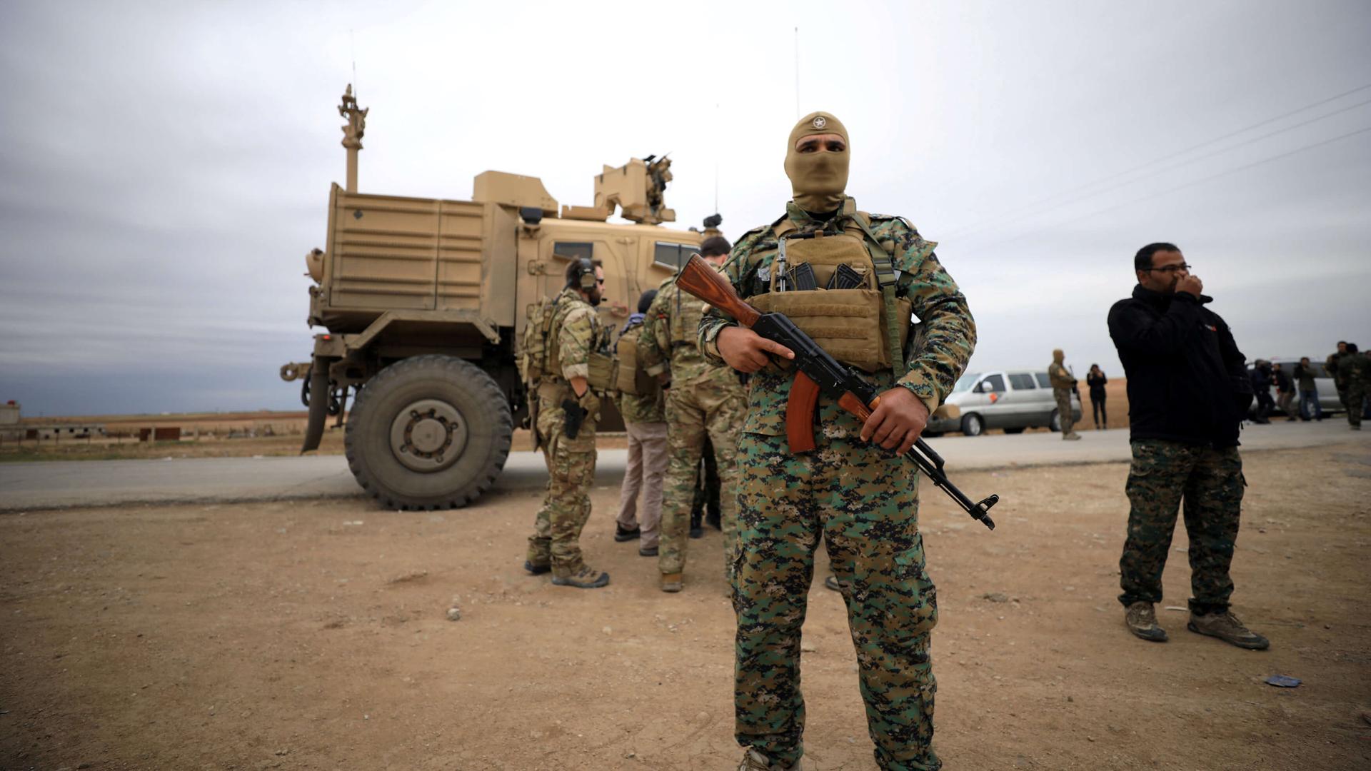 A masked Syria fighter stands in front of an American tank and some troops. 