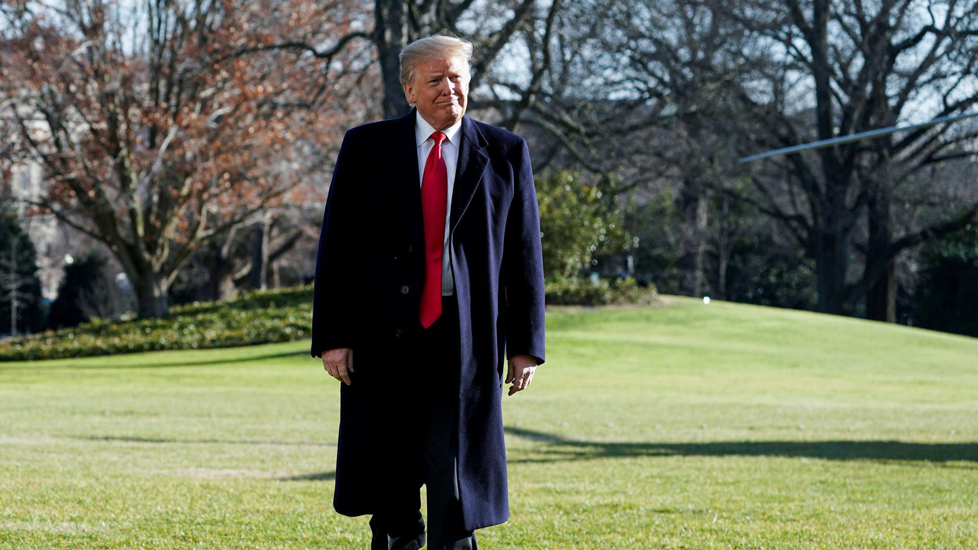 US President Donald Trump is shown walking on a grassy lawn with a dark overcoat on.