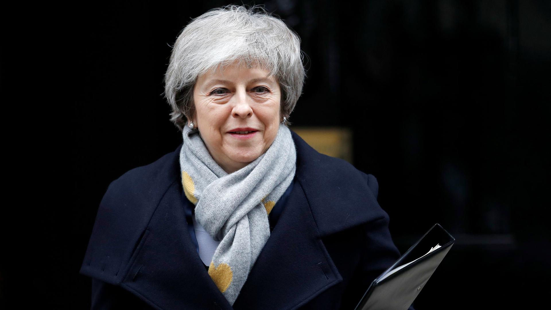 Britain's Prime Minister Theresa May is shown wearing a grey scarf and blue overcoat with a binder in her left hand.