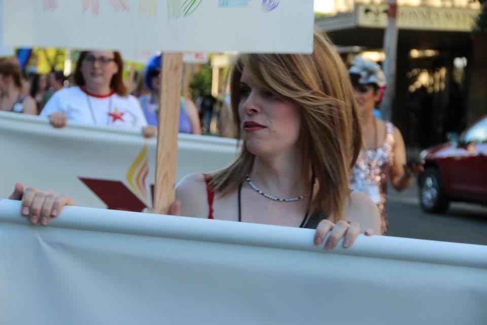 A white woman with medium length light brown hair holds a banner while in a protest march. 
