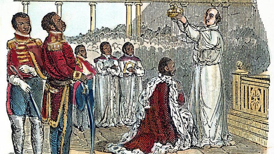 A king in a red robe kneels on one knee as a bishop places a crown on his head