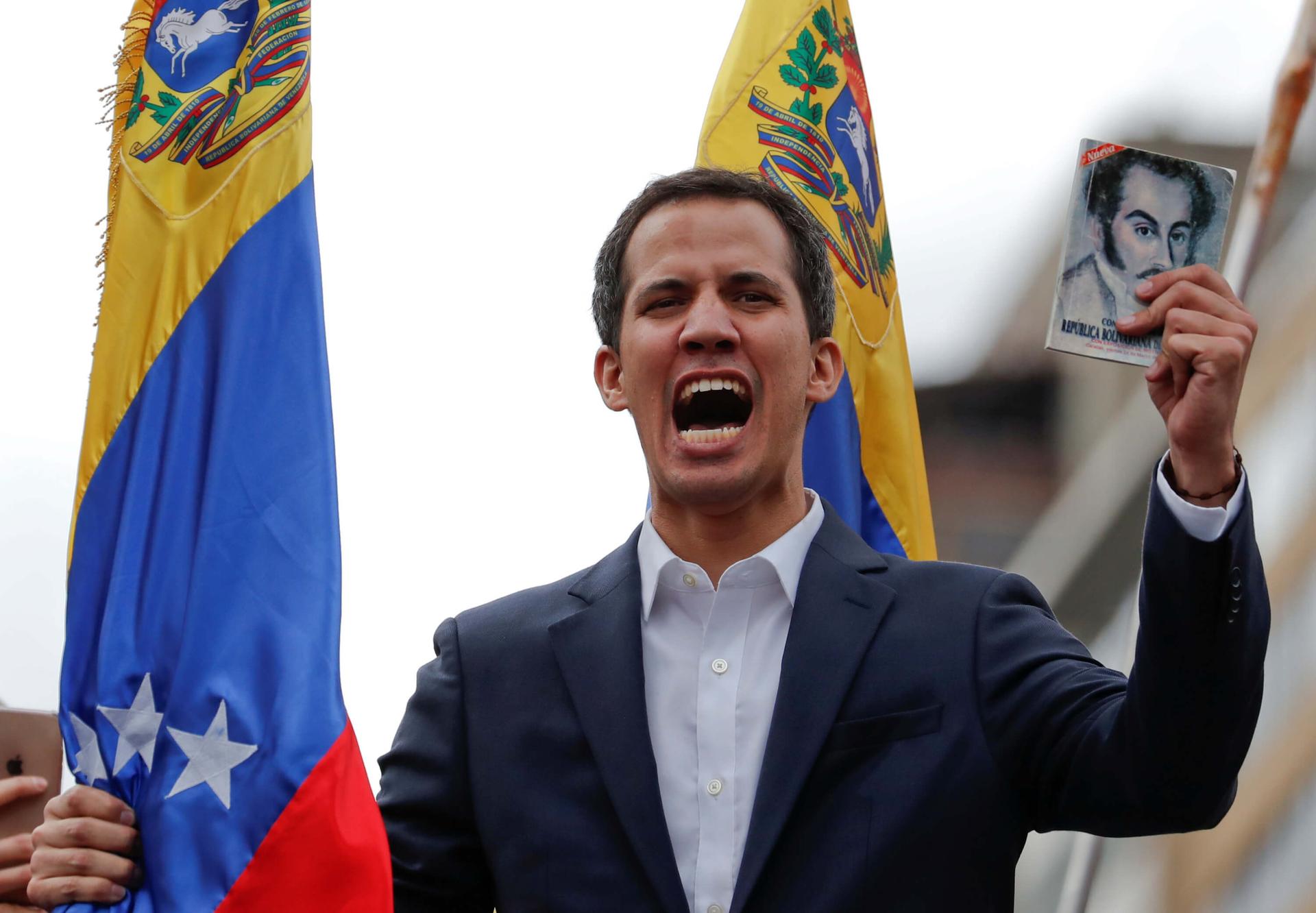 Juan Guaidó  holds a copy of Venezuelan constitution and shouts, in front of the Venezuelan flag