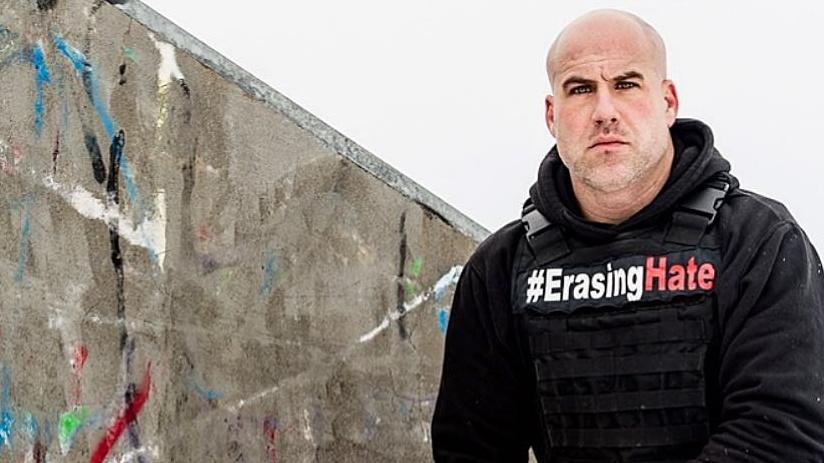 a bald man crouches down next to a graffiti-covered stone. he wears a vest that says "erase the hate"