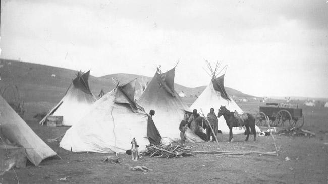 Black and white image of several people and a horse standing in front of teepees. A dog howls in the mid-foreground. 