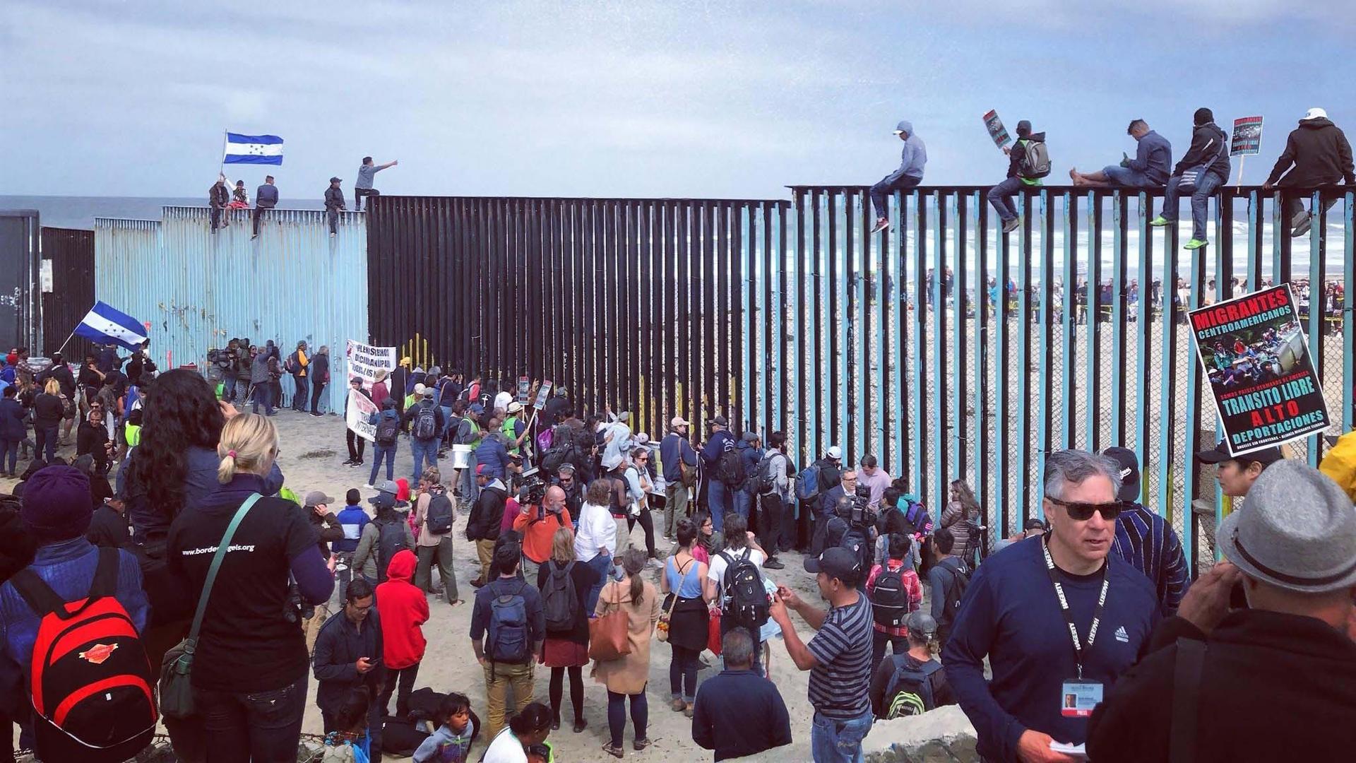 Some people sit on top of a border barrier while a crowd gathers on the ground.