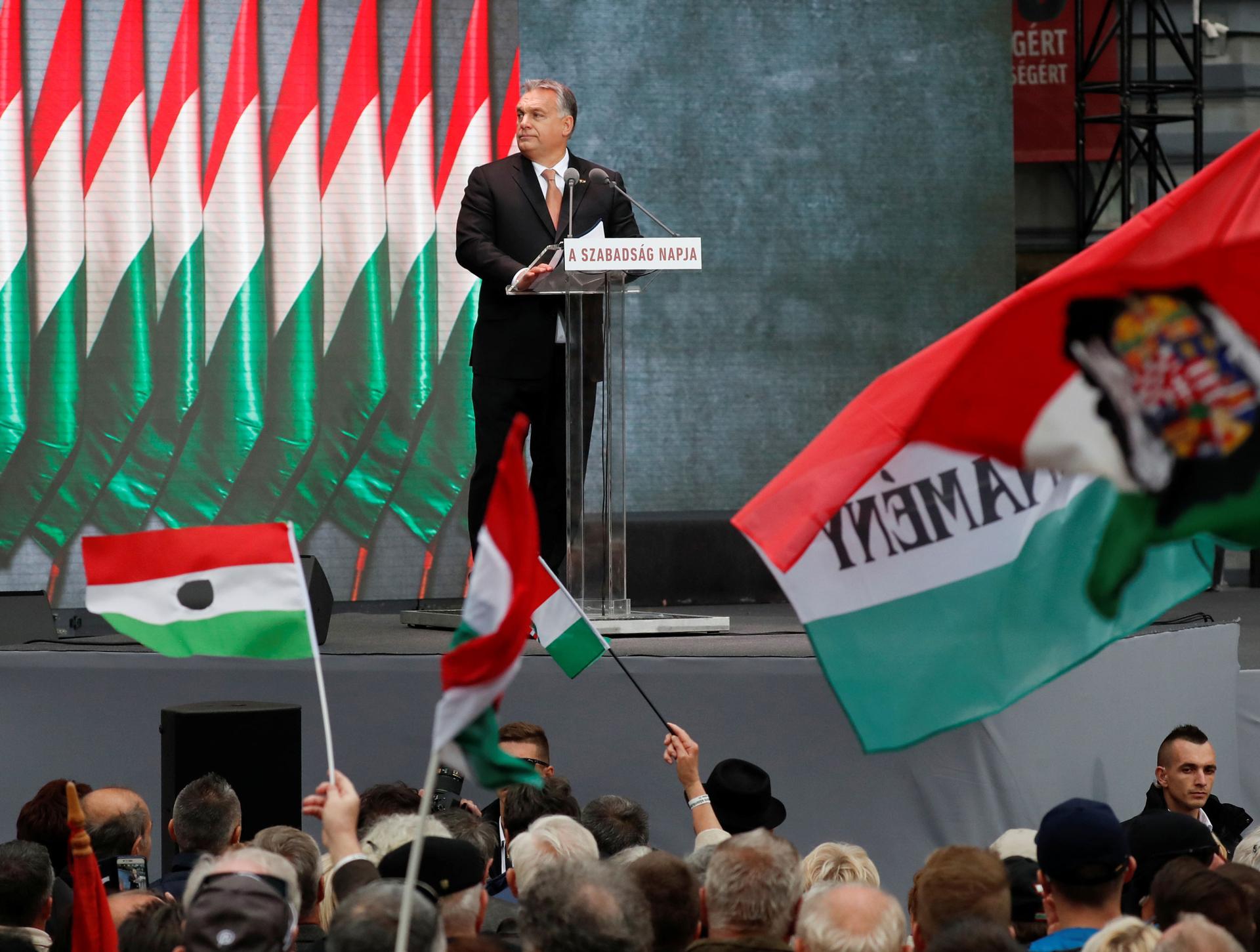 Hungarian Prime Minister Viktor Orbán stands at a podium on a stage above a crowd of people, many of whom are holding Hungarian flags.