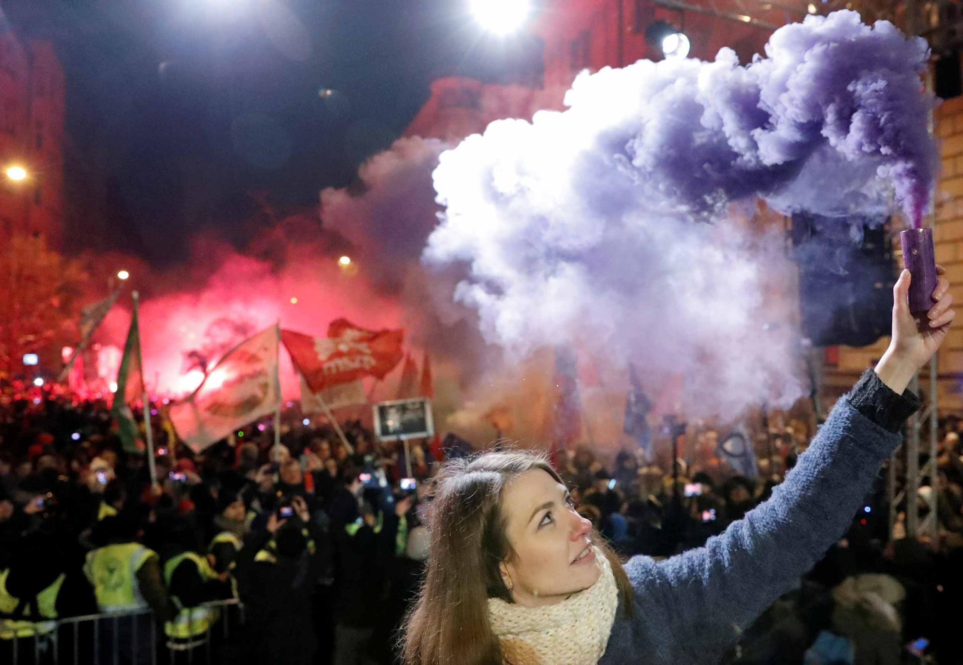 A woman olds a purple flare up the air. Behind her, police clash with protestors in the street.