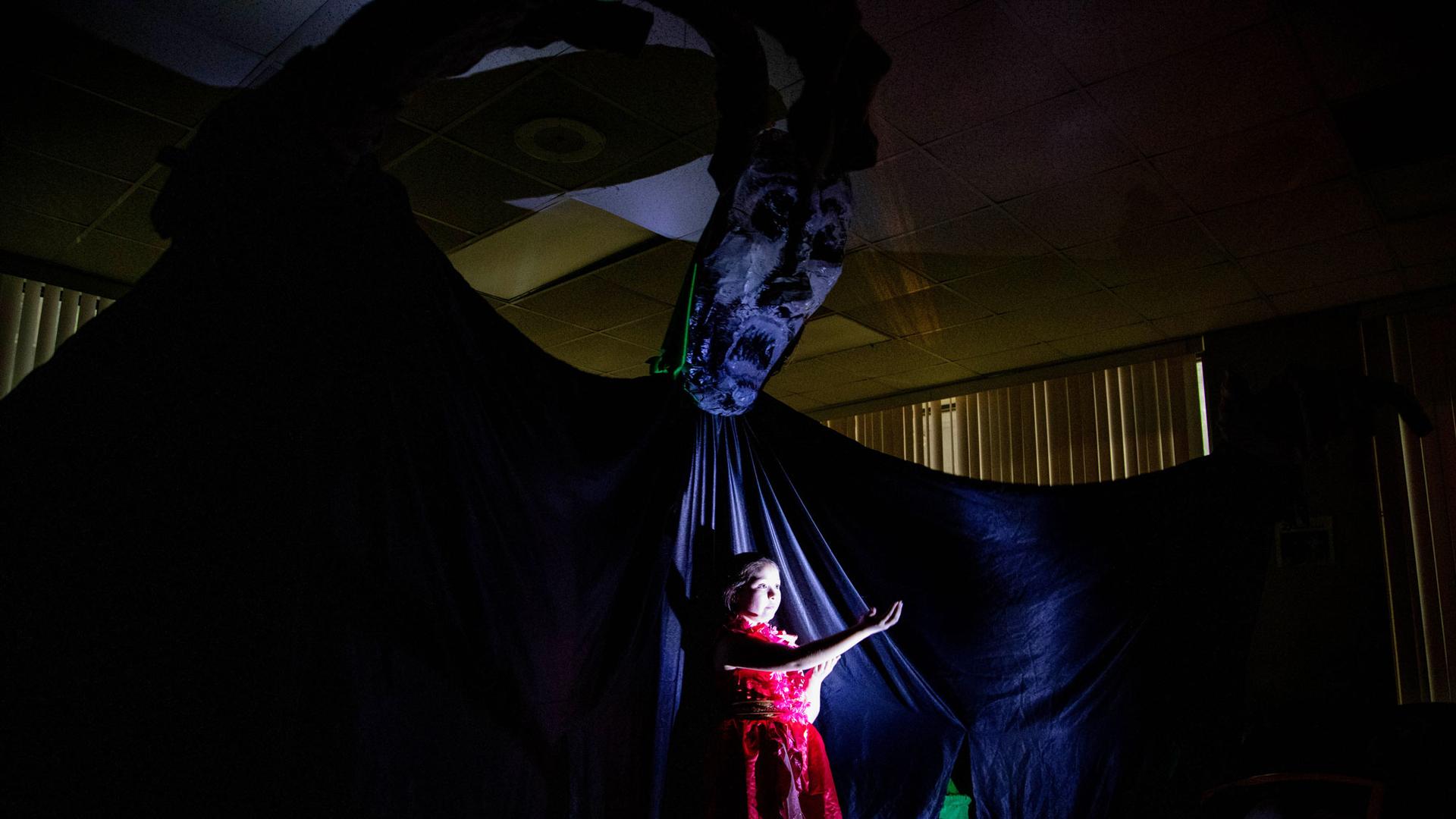 A young girl is lit up on a stage with an enormous dark monster behind her. 