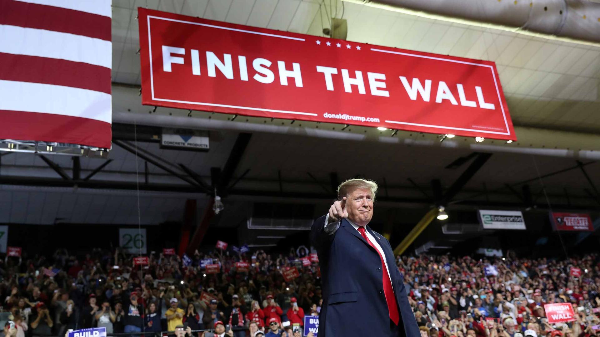 Donald Trump stands in an arena with a banner overhead that reads "Finish the wall."