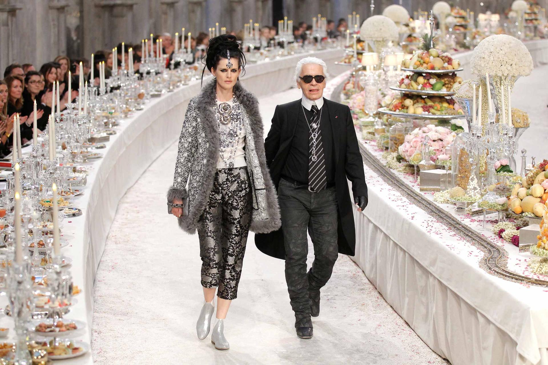 British model Stella Tennant walks with designer Karl Lagerfeld through a runway where people are seated as if for a giant banquet, with candles, crystal and piles of colorful food.